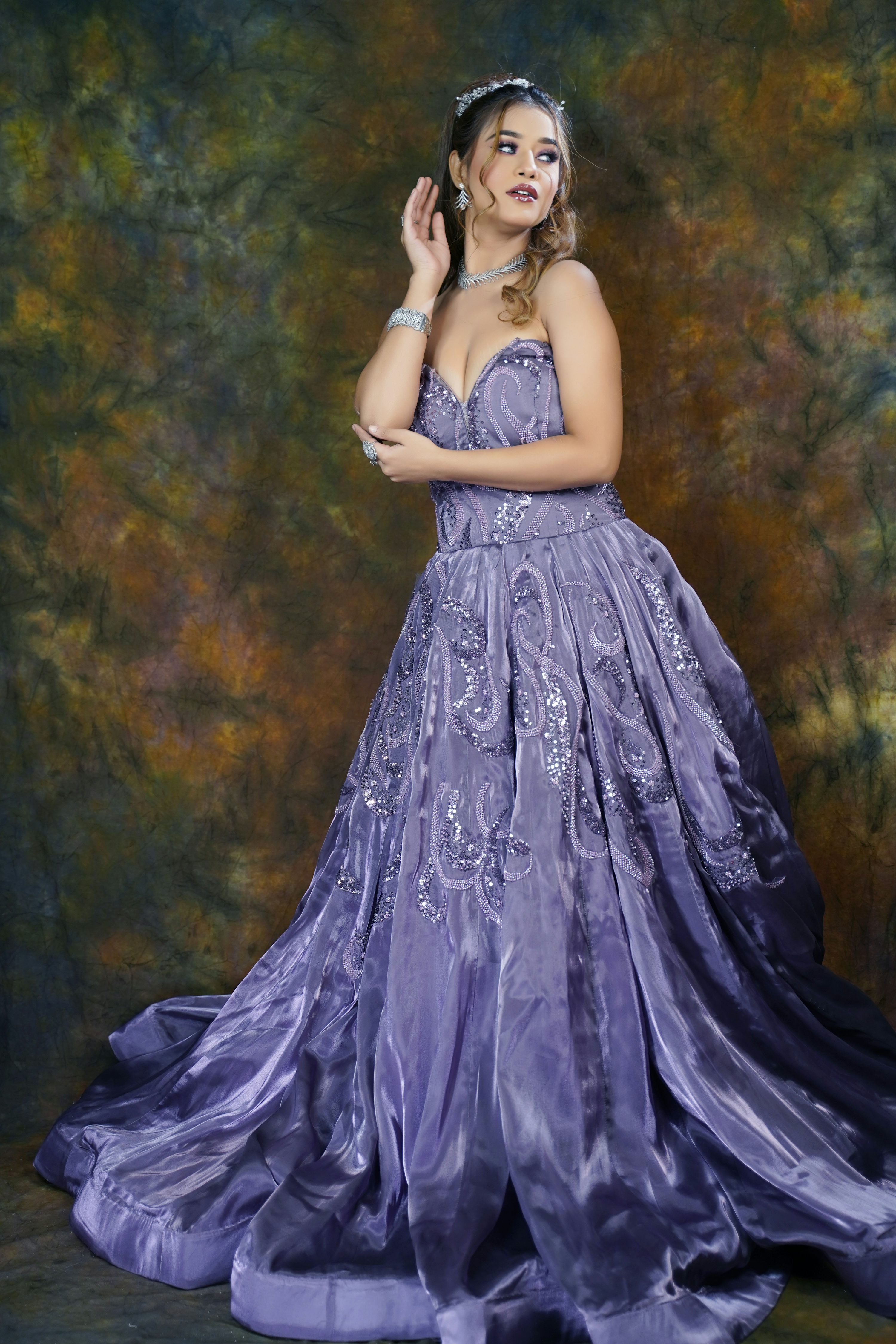 a fulllength portrait of a woman in a beautiful purple ballgown. the gown is strapless with a sweetheart neckline and features intricate sequin and embroidery details. the model is posing gracefully, with one hand gently touching her ear. she wears jewelry including a tiara, earrings, necklace, and bracelet. the background is a textured, dark purple and gold backdrop. the overall mood is elegant, sophisticated, and romantic.