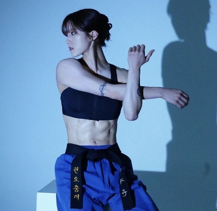 a woman with a black belt and blue pants is stretching her arms and back. she is athletic and toned and has a tattoo of a barbed wire on her arm. she is posing in a studio with light background.