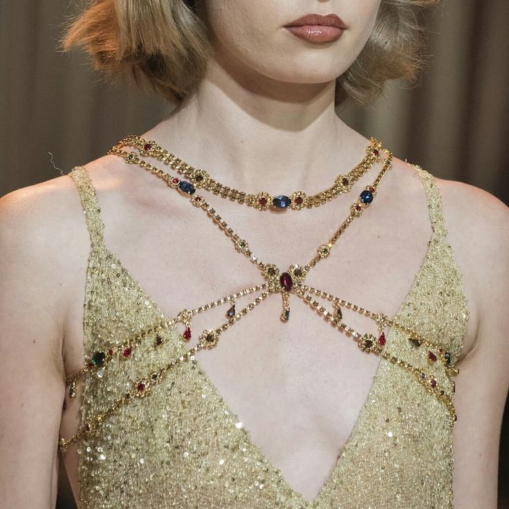 closeup of a woman wearing a golden sequined dress and elaborate gold jewelry. she has a layered necklace and a body chain adorned with rhinestones and colorful gemstones. her blonde hair is styled short, and she wears lipstick. the focus is on the jewelry and dress.