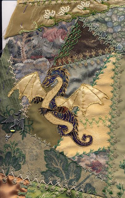 a detailed closeup of a handcrafted textile art piece featuring an embroidered dragon. the dragon is primarily blue and gold, with intricate details. the piece is made of various fabric patches with different textures and patterns, including floral designs, leaves, and beadwork embellishments. the stitching adds to the ornate and decorative appearance of the artwork.