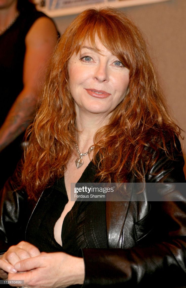 a close up portrait of a beautiful redhead woman in a black leather jacket, she is smiling and looking at the camera, her red hair is curly and flowing down her shoulders. she is wearing a black shirt and a necklace with a heart pendant. the woman is sitting and appears to be in a studio setting.