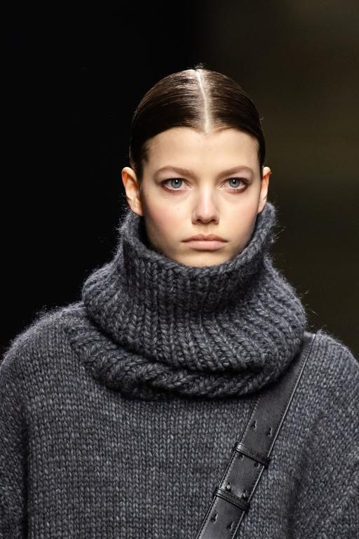 a young woman with dark hair and gray sweater poses for a studio portrait. she is wearing a gray turtleneck sweater with a matching knit scarf. she is looking at the camera with a serious expression. the background is dark and blurred.