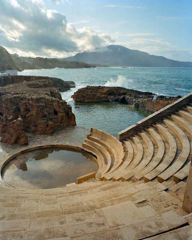 this image showcases a picturesque coastal scene featuring an amphitheater built into the landscape. the amphitheater's curved stone steps lead down to a circular pool, reflecting the sky and clouds above. beyond the amphitheater, rocky formations extend into the ocean, with waves crashing against them. in the distance, a mountain rises into the sky, partially obscured by clouds, completing the stunning seascape.