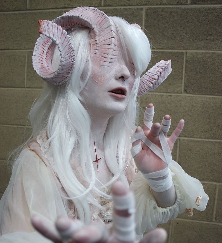 a young woman with white hair and pale skin, wearing a white dress and horns, has her eyes covered with a bandage.  she is wearing a white dress with lace trim.  her hands are bandaged. her face is heavily made up with white paint and dark circles around her eyes.  her horns are large and ornate.