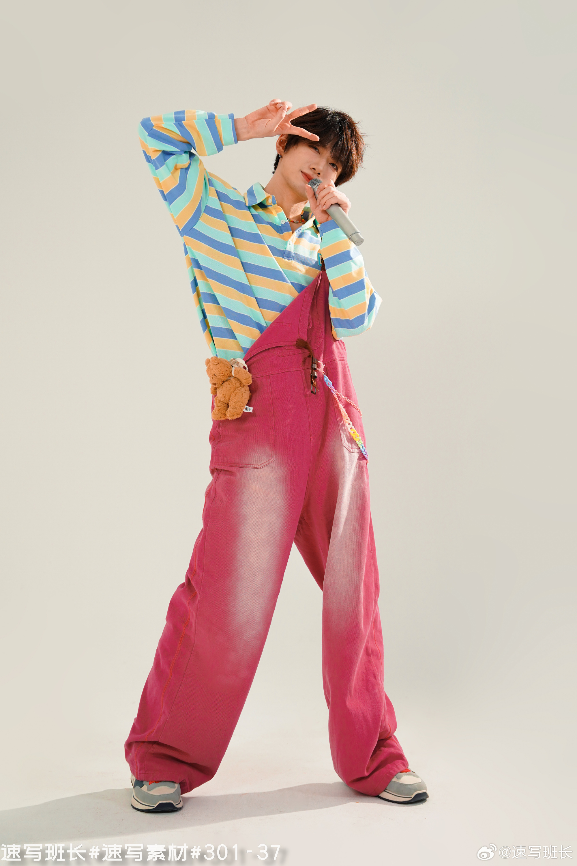 a young asian man in a playful outfit, consisting of a colorful striped shirt and pink overalls, is posing against a white background. he's holding a microphone in one hand and making a peace sign with the other. a small teddy bear is attached to his overalls. the lighting is bright and artificial, highlighting the vibrant colors of his clothing.