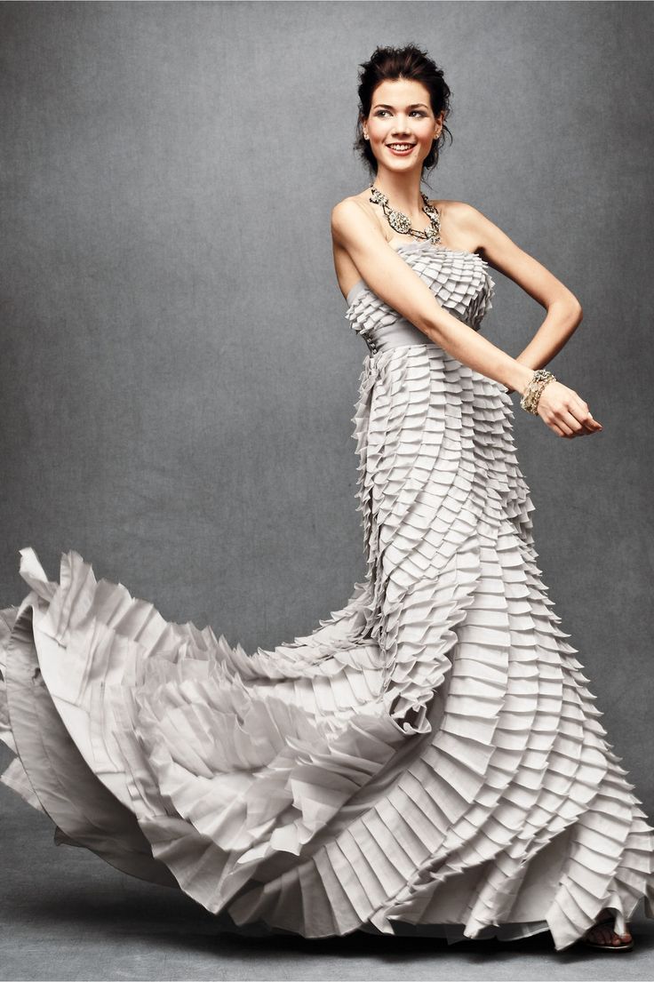 a beautiful woman in a flowing gray evening dress stands against a white background. the dress is a sophisticated design, with ruffles, and a floorlength hem. the woman is smiling, and her hair is pulled back, she has elegant jewelry and make up. she is holding her arms out in front of her, with her hands placed together, looking to the side.