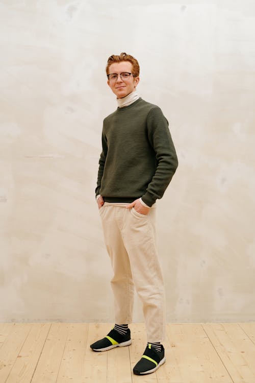 a fullbody shot of a redhead man with glasses standing indoors. he is wearing a white turtleneck, an olive green sweater, beige pants, black and green athletic shoes, and striped socks. he has his hands in his pockets and is smiling. the background is a lightcolored wall and a wooden floor.