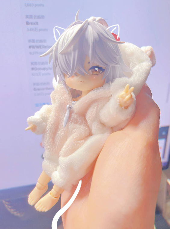 a small, white, fluffy anime doll is being held in someone's hand. the doll is wearing a fuzzy white and pink jacket with a hood, and has cat ears. she is making a peace sign with her left hand. the doll is so small, that she is sitting in the palm of someone's hand.
