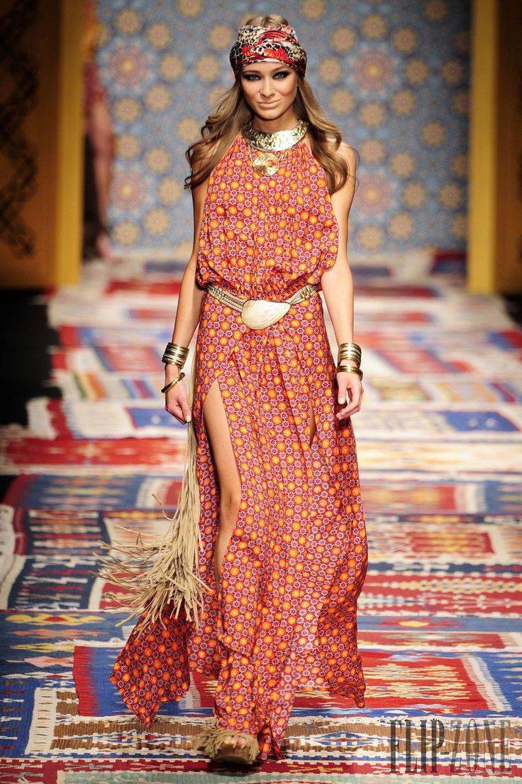 a model in a red and orange floral print dress with a side slit, walking the runway. she has a gold head band on her head, a gold necklace, and a gold belt with a large buckle. she is also wearing gold bangles on her arms, sandals, and has a large, fringed scarf hanging from her dress.