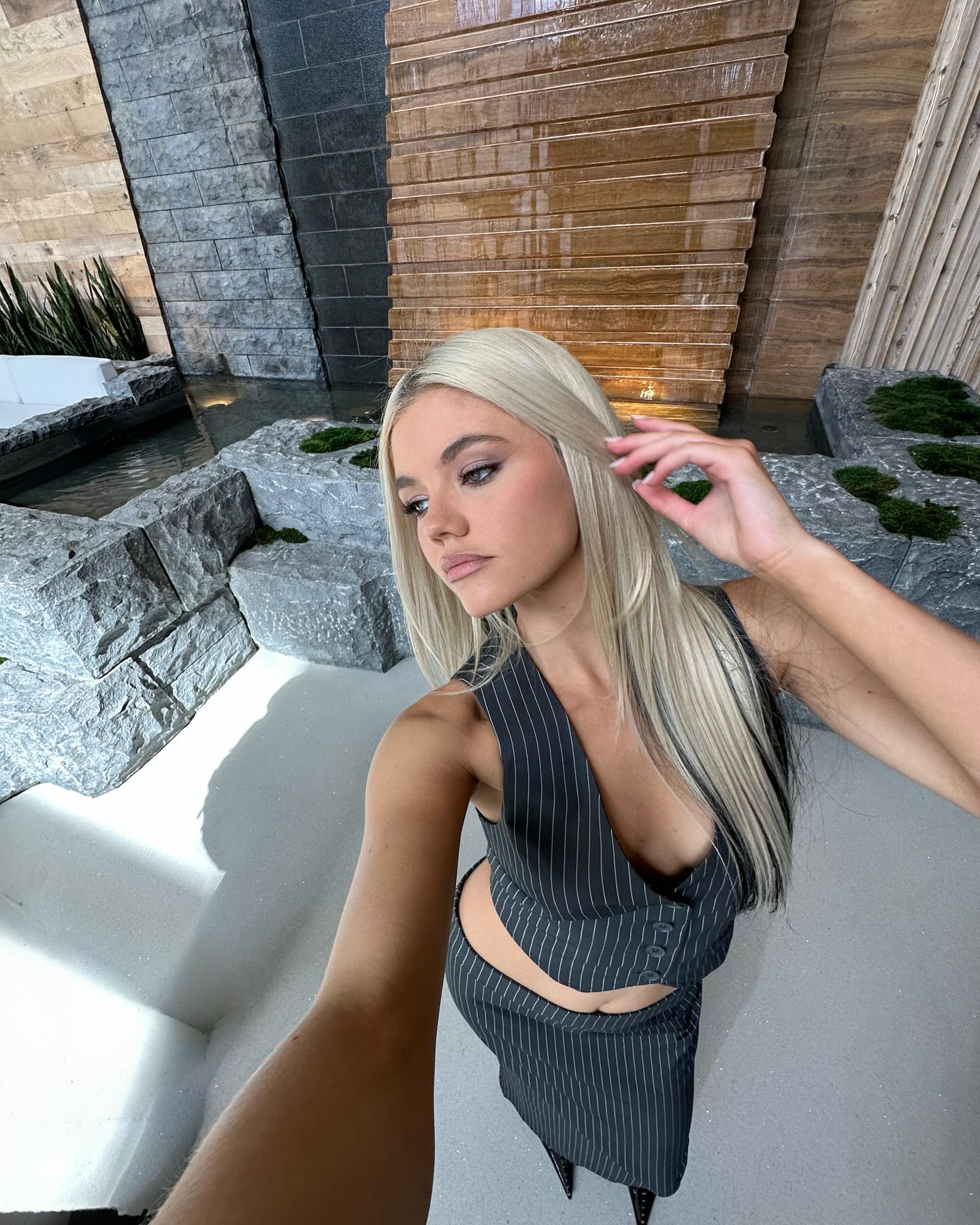 a blonde woman wearing a pinstripe suit takes a selfie in front of an indoor waterfall with rocks and a water feature. she is posing and adjusting her hair.