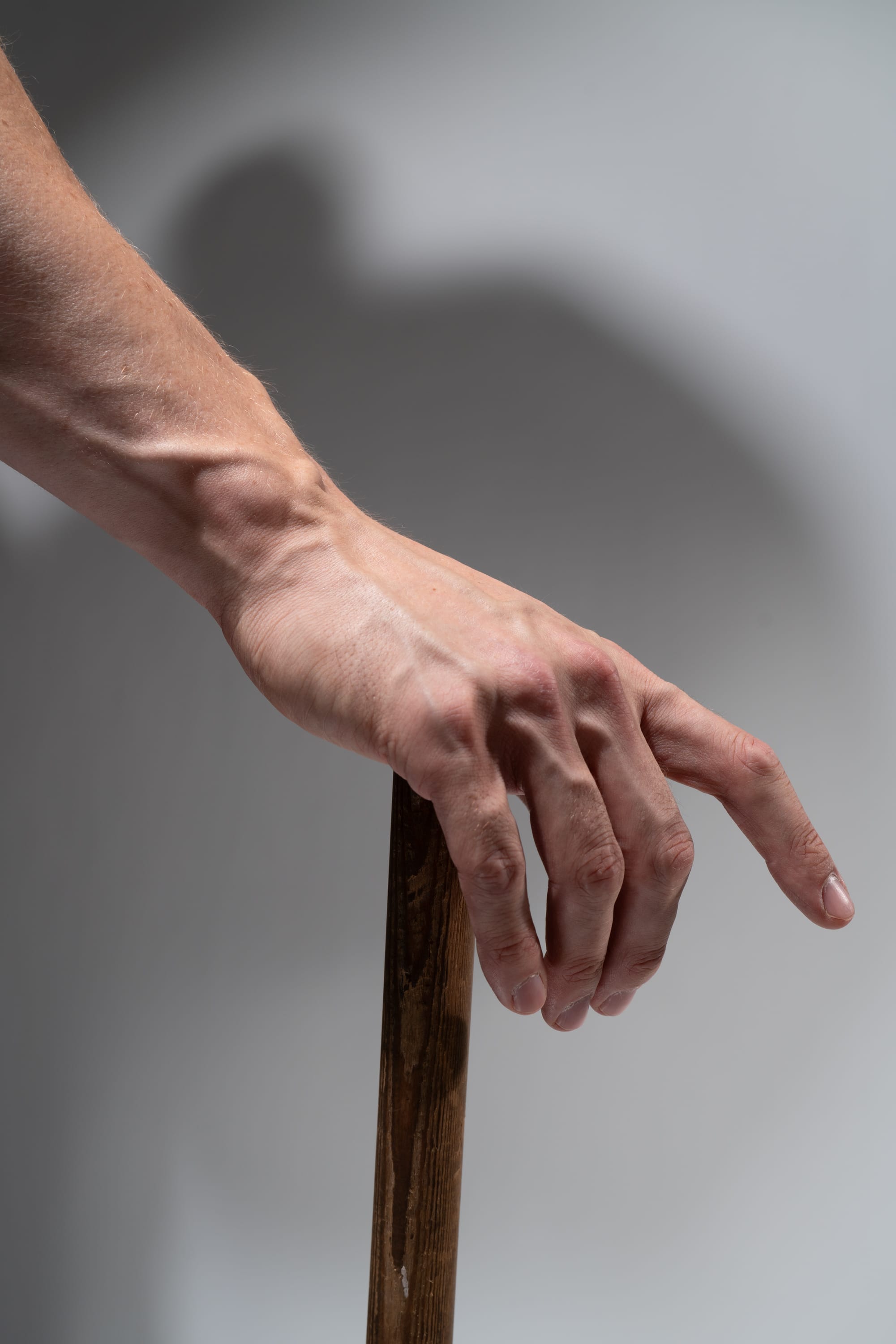a closeup shot of a man's hand gripping a wooden walking stick. the hand is veiny and the light casts a subtle shadow on the background. the image evokes themes of aging, disability, and reliance on assistive devices.