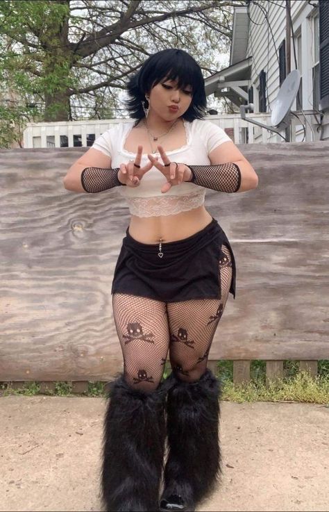 a fullbody shot of a woman in goth fashion standing outside. she is wearing a white crop top with lace trim, a short black skirt, fishnet stockings with a skull pattern, and furry black boots. she has short black hair with bangs and is wearing jewelry. her hands are raised in front of her, forming a vshape with her fingers. the background includes a wooden fence, grass, and a house.