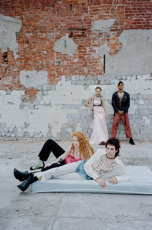 the photograph captures a group of young models, exhibiting a punk or grunge aesthetic, posing against a backdrop of a weathered brick wall. the scene is gritty and urban. two female models are positioned standing and lying, one in a light pink dress and the other in a pink top and dark pants, alongside two male models. one male model sports a black leather jacket and plaid pants, and the other is dressed in a white shirt. the models are arranged on and around a light blue mattress, lending a sense of casualness and rebellion against typical fashion photography settings. the overall mood is edgy and alternative, reminiscent of 90s youth culture.