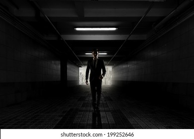 a man in a suit is walking through a dark tunnel towards the light at the end. the tunnel is dimly lit with overhead lights, and the man's silhouette is prominent against the light. he is walking towards the viewer with a determined stride.