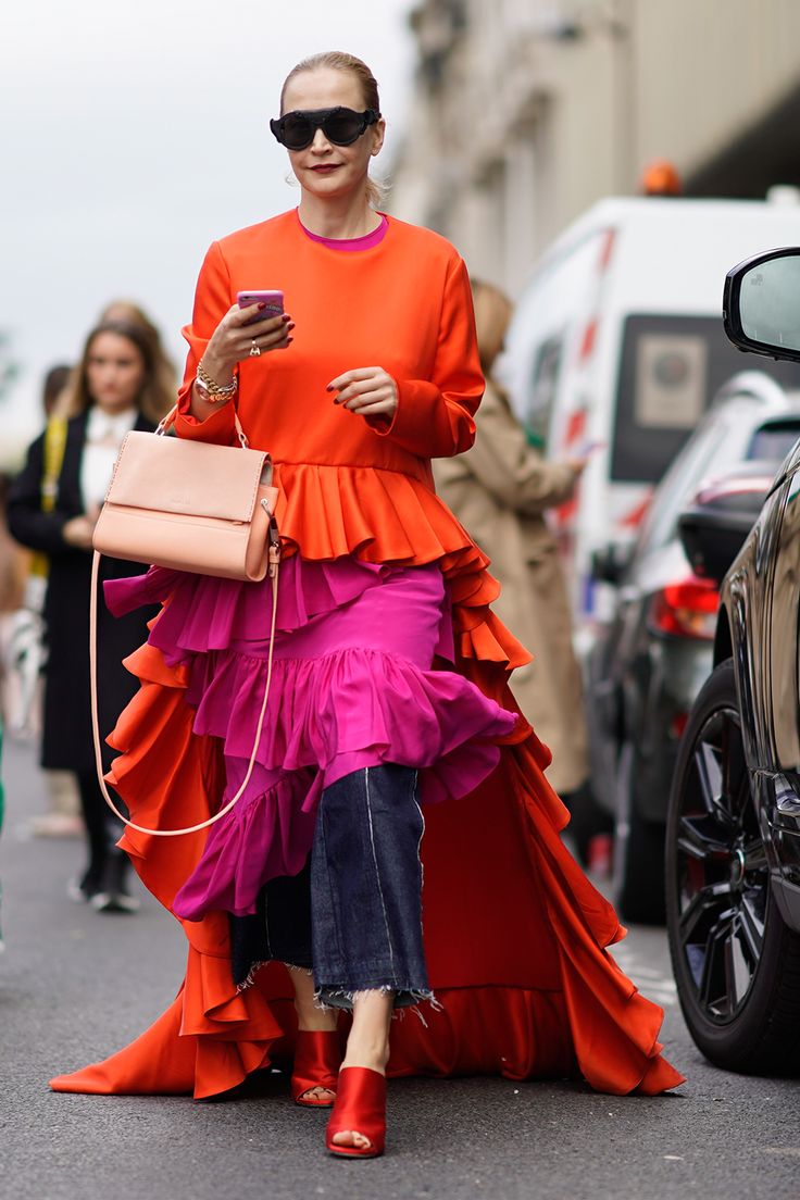 a woman stands on a city street holding a cell phone. she is wearing oversized sunglasses, a bright orange top and a pink top underneath, an orange and pink ruffled skirt with a long orange train, frayed blue jeans, and red heeled shoes. she is also wearing jewelry and carrying a pink purse. cars and people are blurred in the background.