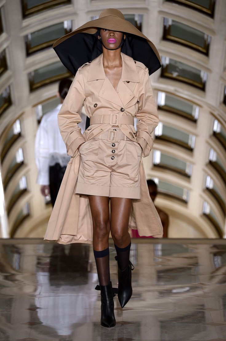 a fashion model walks the runway during a fashion show. she is wearing a beige trench coat with matching shorts, black boots, and sheer black socks. she also sports a large, widebrimmed beige hat. the background is a grand, elegant hallway with arched features. the overall style is elegant and sophisticated, with a focus on high fashion.