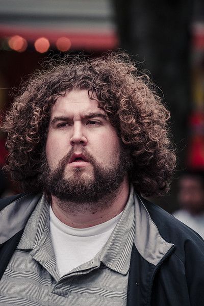 a portrait of a man with curly brown hair and a beard, wearing a white shirt and a black jacket. the background is blurred, suggesting an outdoor setting.