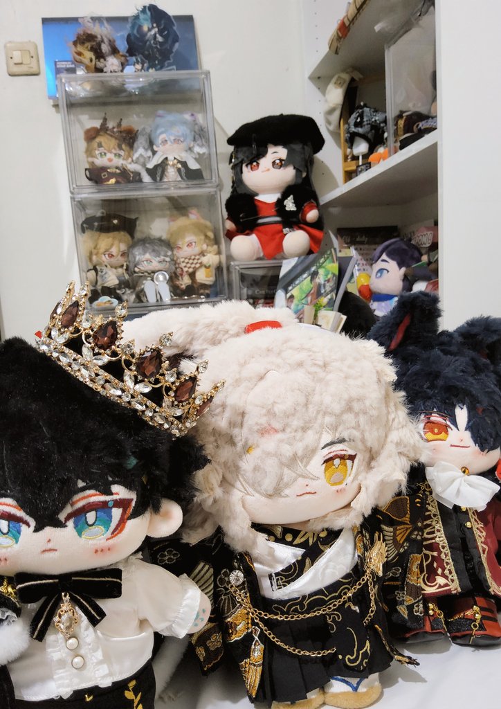 a collection of anime plush toys are displayed on a shelf. they are all dressed in different outfits and are looking at the camera. there are also other items on the shelf, such as figurines and a box of other plush toys. the plush toys are all very cute and cuddly.