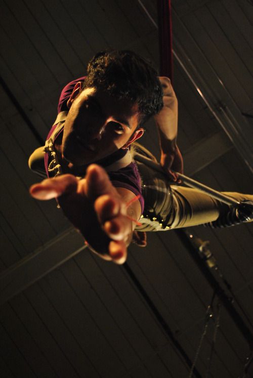 a performer hangs upside down from an aerial silk, suspended in midair. their body is contorted in a challenging pose, showcasing impressive flexibility and strength. the performer's focus and determination are evident as they hold this aerial position. the dim lighting adds to the dramatic effect of the image.