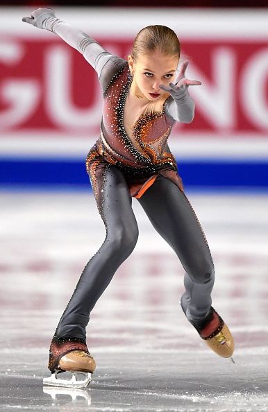 a female figure skater is performing a move on the ice. she is wearing a black and orange costume, and her left skate is off the ice while she is in the air. the background is blurry and the word "gunc" is seen in the background.