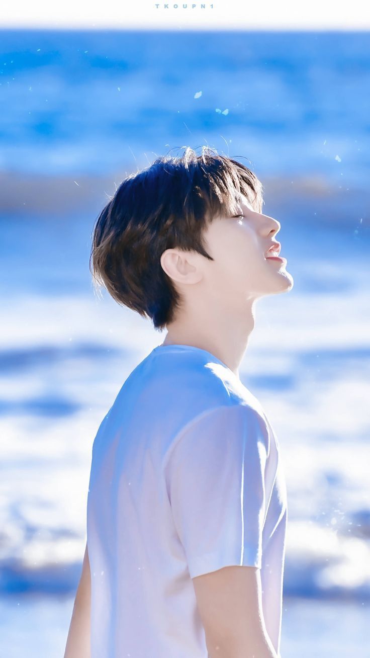 a young man with short dark hair, wearing a white tshirt, is standing on a beach, facing towards the right with his head tilted back and eyes closed, as if enjoying the warmth of the sun. the background features a clear blue sky and sparkling ocean waves. the overall ambiance is bright, cheerful, and serene.