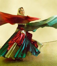 a woman belly dances in a colorful dress and flowing fabric. she is spinning, and the skirt of her dress is flared out around her. the fabric in her hands is flowing out to the sides. she is smiling.