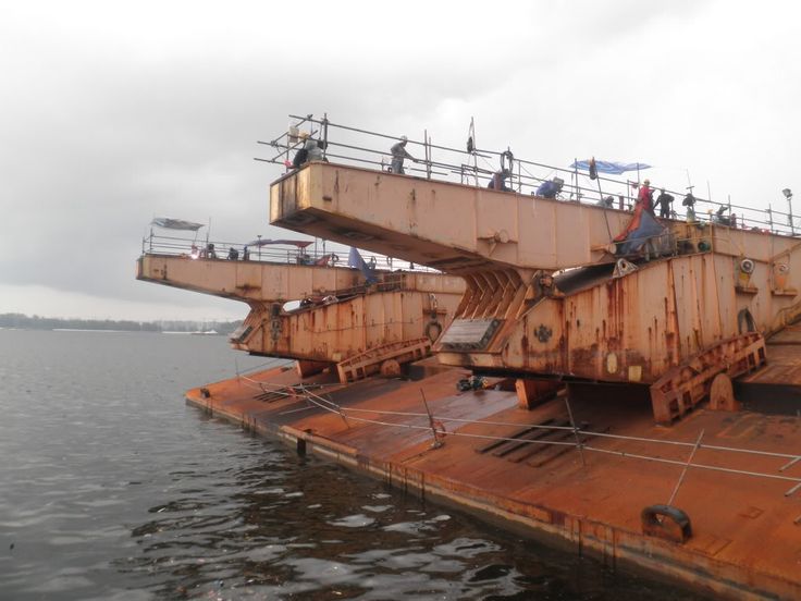 the image shows a large, rusty barge or floating platform in a river.  multiple large, rusty metal structures, possibly parts of a crane or lifting mechanism, are situated on the barge.  numerous workers can be seen on the scaffolding surrounding these structures, suggesting the barge is undergoing dismantling or demolition. the overall appearance is one of industrial decay and salvage operations. the sky is overcast.
