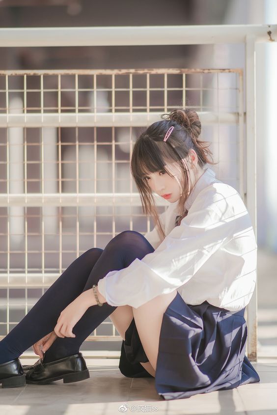 a young woman in a school uniform is sitting on the ground in front of a metal fence. she is wearing a white shirt, navy blue skirt, black stockings, and black loafers. her hair is styled in a messy bun with bangs and a pink hair clip. she has a bracelet on her left wrist and is looking down.