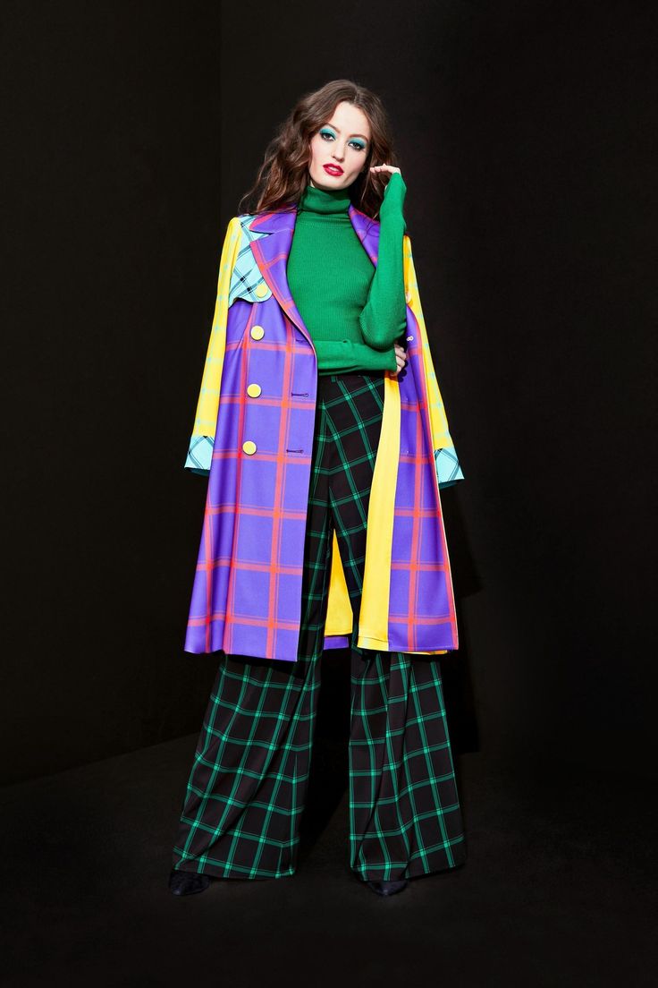 a fulllength shot of a female fashion model posing against a black background. she is wearing a vibrant, multicolored coat with a plaid pattern in shades of purple, yellow, and orange. the coat is a long, loose fit and has large, bold checkered patterns. underneath the coat, she wears a bright green turtleneck sweater and widelegged pants with a green and black plaid pattern. her hair is styled in loose waves and she has a bold makeup look. the overall style is a mix of playful and sophisticated. the image looks like it's from a professional fashion photoshoot.