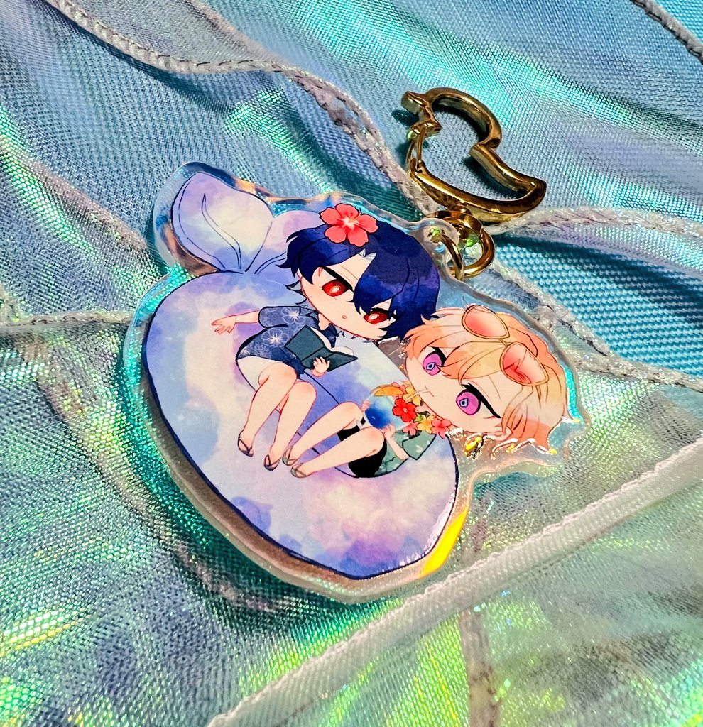 a close up of an anime style keychain featuring two boys on a blue and pink donut float in the water. the boys are both wearing swimsuits and smiling. the keychain is made of clear plastic with a gold ring attached. the background is a blue iridescent fabric.
