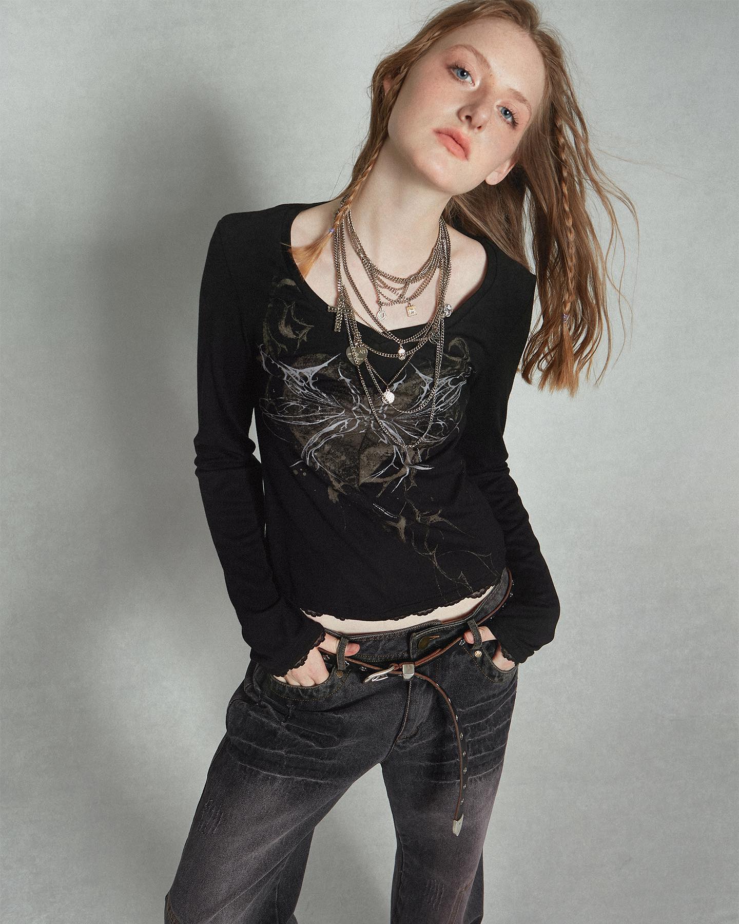 a young woman with long braided hair is posing in a studio shot against a light grey background. she is wearing a black longsleeved shirt with a graphic design, dark wash jeans, and a brown belt. she is also wearing multiple layered necklaces. her style is casual yet edgy, with a slightly goth or grunge influence.  the image is welllit and shows the details of her clothing and accessories.