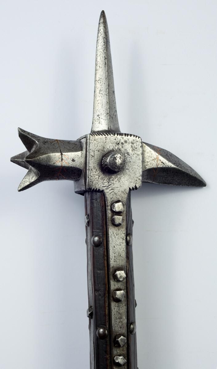 closeup shot of a medieval war hammer. the hammerhead is made of steel, featuring a prominent spike at the top and a hammerhead with multiple points on one side and a curved beak on the other. the head is attached to a wooden shaft reinforced with steel strips and rivets. the background is plain white.