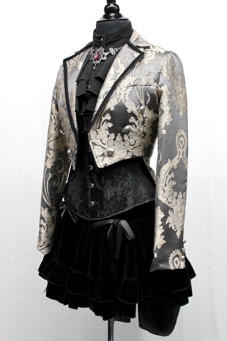 a closeup image of a black and silver outfit,  a black velvet, ruffled skirt paired with a silver brocade jacket, black corset, and black velvet bow. the jacket has silver trim, silver buttons, and silver jewelry. the outfit is styled to look like a gothic or steampunk fashion.  it's displayed on a mannequin in a fashion boutique, with white background.  the outfit gives a sense of luxury, formal wear, and special occasion.  the image captures the details of the clothing and the styling, making it look visually appealing.