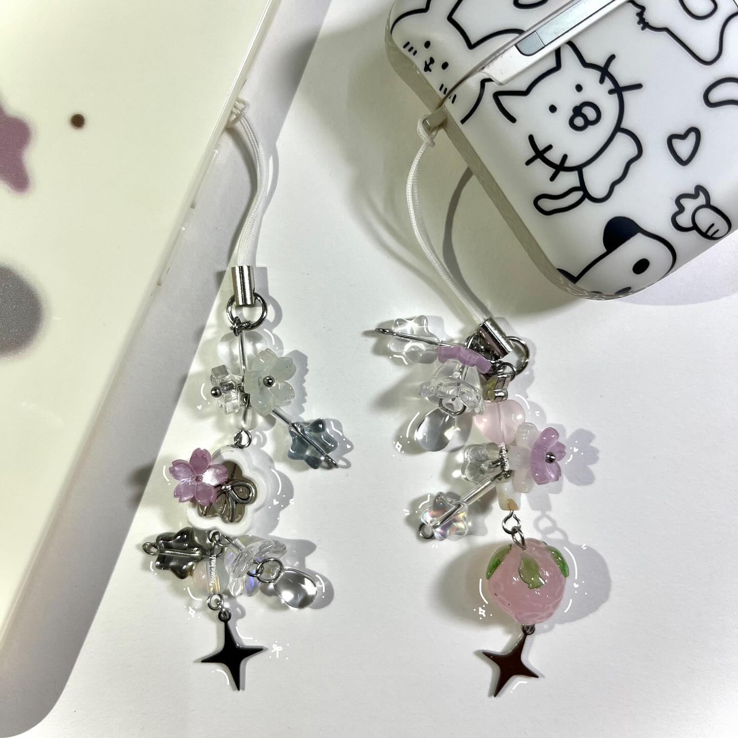 the image shows two devices with charms attached. on the left is a white phone in a clear case. a phone charm is hanging off it with multiple beads, metal charms, and a star. on the right is an airpods case with a cartoon cat and dog drawing on it, along with a heart. a similar beaded charm is hanging from the case.