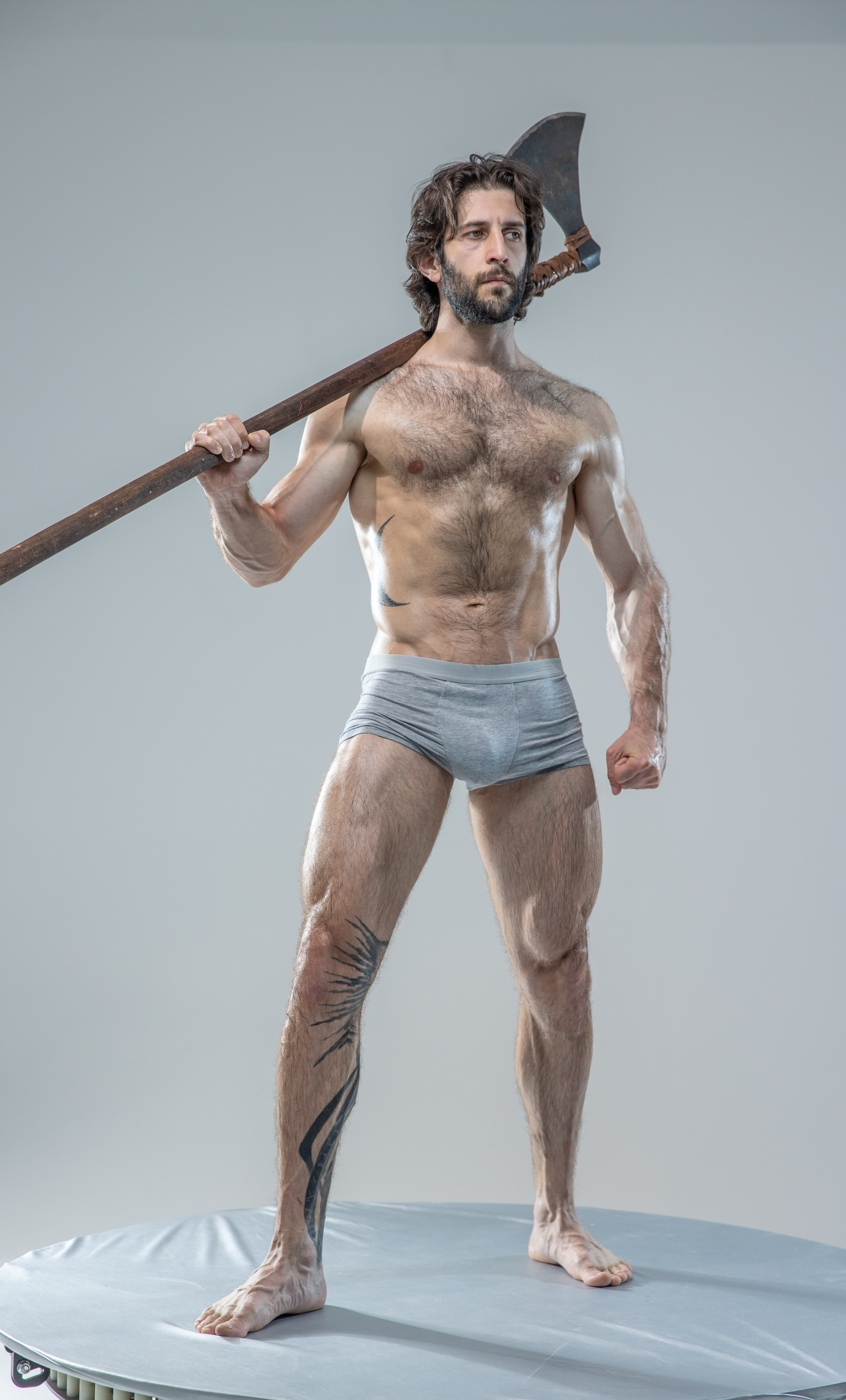 a muscular man with long, dark, curly hair and beard is standing on a round platform against a light grey background. he is wearing grey underwear and is holding a large, doubleheaded axe over his right shoulder. he has tattoos on his left leg and has a serious expression on his face. his body is toned and he appears strong and powerful.