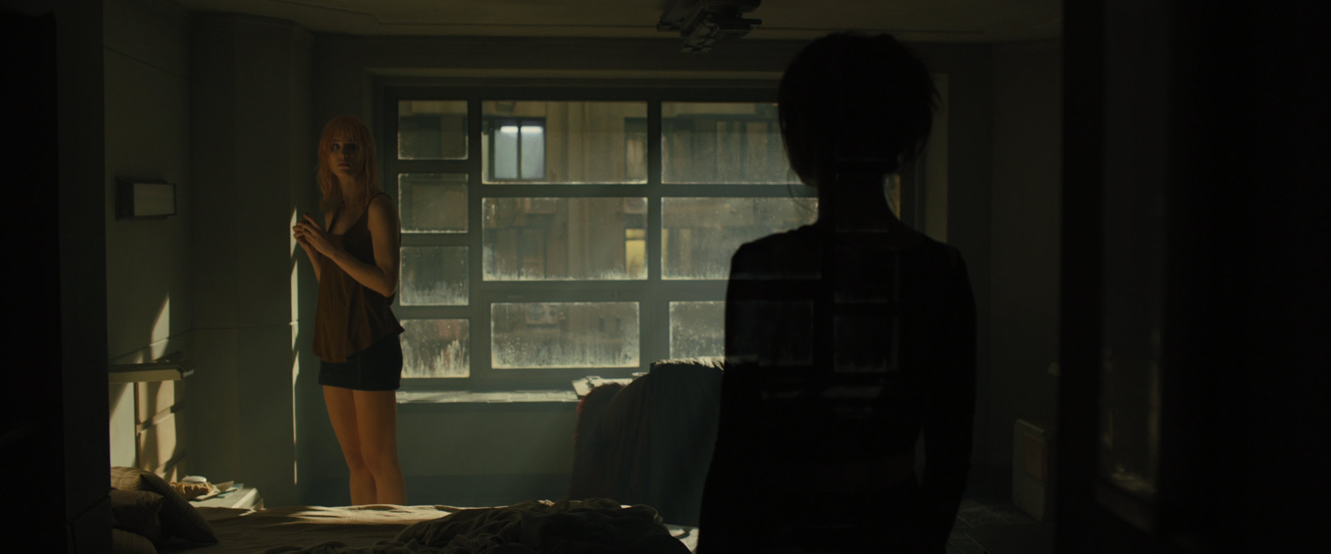 a woman with short, pink hair stands by a window in a dimly lit room. she is wearing a brown tank top and black shorts. the window is grimy and overlooks a city street. the room is sparsely furnished with a bed in the foreground. another person, only visible as a silhouette, stands in the foreground with their back to the camera. the overall atmosphere of the image is moody and atmospheric.