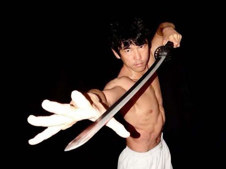 a shirtless man with dark hair is wielding a katana in front of a black background. he is holding the sword in his right hand and extending his left hand toward the viewer. he is wearing white pants.