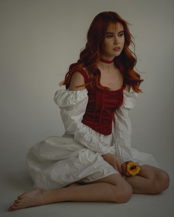 a young woman with long red hair is sitting on the floor, looking down. she is wearing a white dress with a red bodice and holding a peach. the background is a light gray wall. the woman's expression is serious and thoughtful.