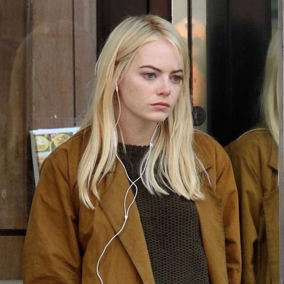 a photo of emma stone with blonde hair wearing earphones and a brown coat over a green sweater, looking pensive and melancholic. she's outdoors with a reflection of herself in the background. she is wearing natural makeup.