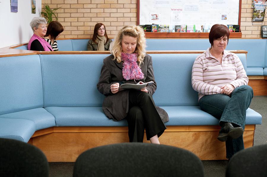 a waiting room with a blue couch with five women sitting on it. three women are on the same couch in the foreground and the other two are on a couch in the background. one of the women in the foreground is reading a magazine and another is looking down. the women in the background are looking in different directions. the room is decorated with a brick wall, a bookshelf, and a wooden floor.