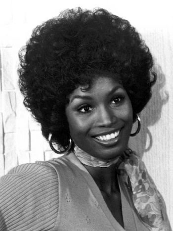 a black and white portrait of a smiling african american woman with afro hair and earrings, wearing a scarf and a knitted sweater, against a plain background.