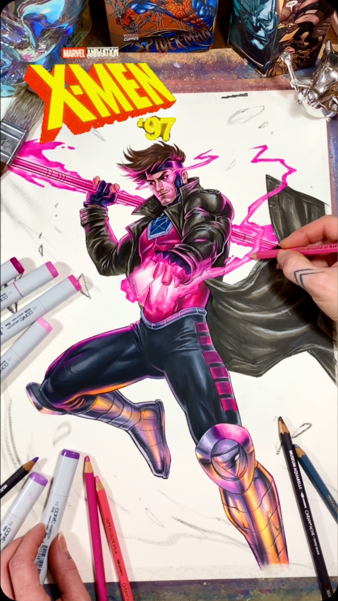 a closeup shot of a drawing of gambit from the xmen, a marvel comics superhero.  he is holding a pink energy blast in his hand, and is surrounded by pink energy bolts. there are various colored pencils and markers on a table in the foreground, along with a small figure in the background.