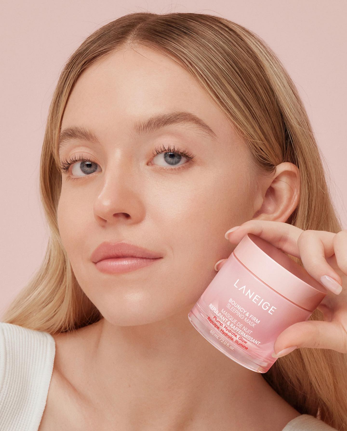 a closeup promotional image features actress sydney sweeney holding a jar of laneige bouncy & firm sleeping mask. she is fairskinned with blonde hair and blue eyes, and the pink skincare product is prominently displayed. the image focuses on the beauty product and the model's fresh, clean look.