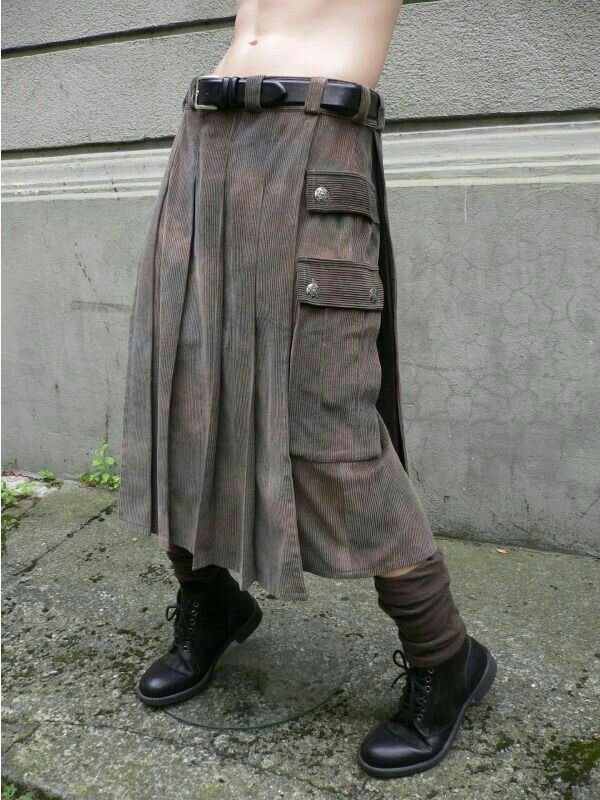 a closeup shot of a mannequin wearing a brown corduroy kilt with cargo pockets and a black belt. the kilt is pleated and asymmetrical, falling longer in the front than in the back. the mannequin also sports black combat boots and brown socks. the background features a grey stone wall and pavement. the overall style is a blend of steampunk, alternative, and grunge fashion elements.