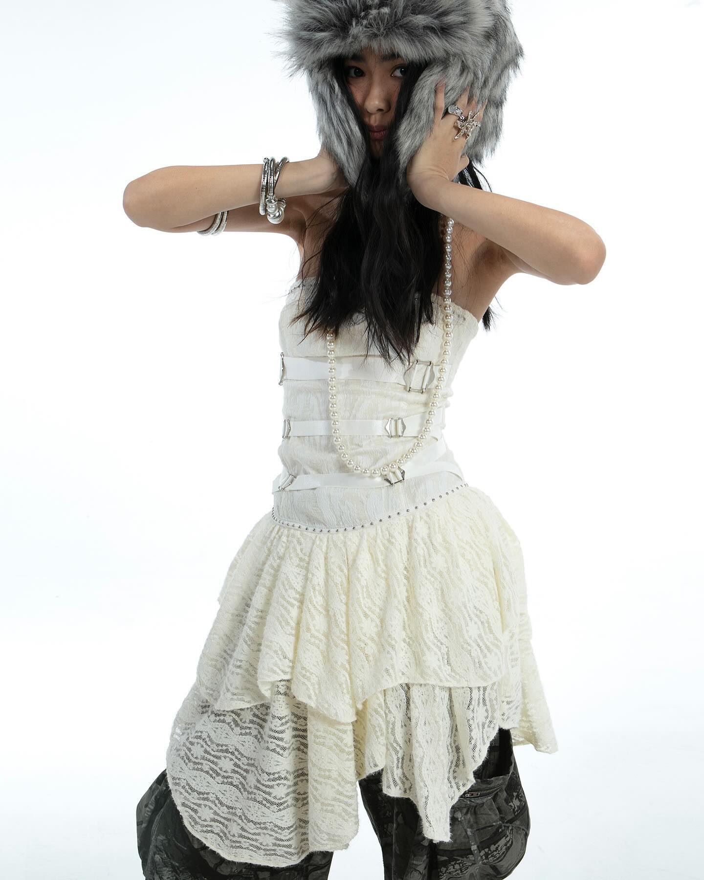 a young woman, possibly a fashion model, is posing against a stark white background. she is wearing a stylish offtheshoulder creamcolored lace dress with multiple layers and an asymmetrical hemline. the dress is cinched at the waist and has a corsetlike bodice with buckles. she is also wearing a grey fur ushanka hat, multiple bracelets, a pearl necklace, and rings. her dark hair is partially concealed by the hat.  the overall style is a blend of elegant and edgy, incorporating both feminine and alternative fashion elements. the image appears to be a professional studio shot for a fashion catalog or editorial.