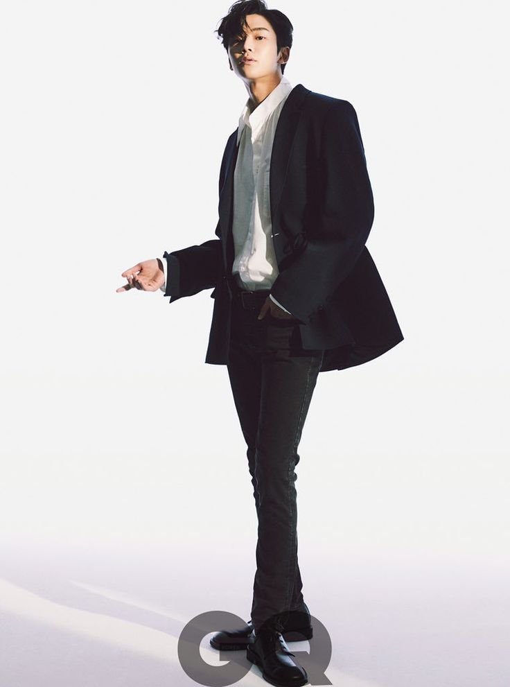 a man, dressed in a black suit, white shirt, and black pants, is posing for a photoshoot in a studio. he is standing with his left leg slightly forward, and his right arm is extended out to his side. he is looking directly at the camera with a serious expression. the background is a white wall, and the floor is a light gray. there is a gq logo on the floor in the foreground.