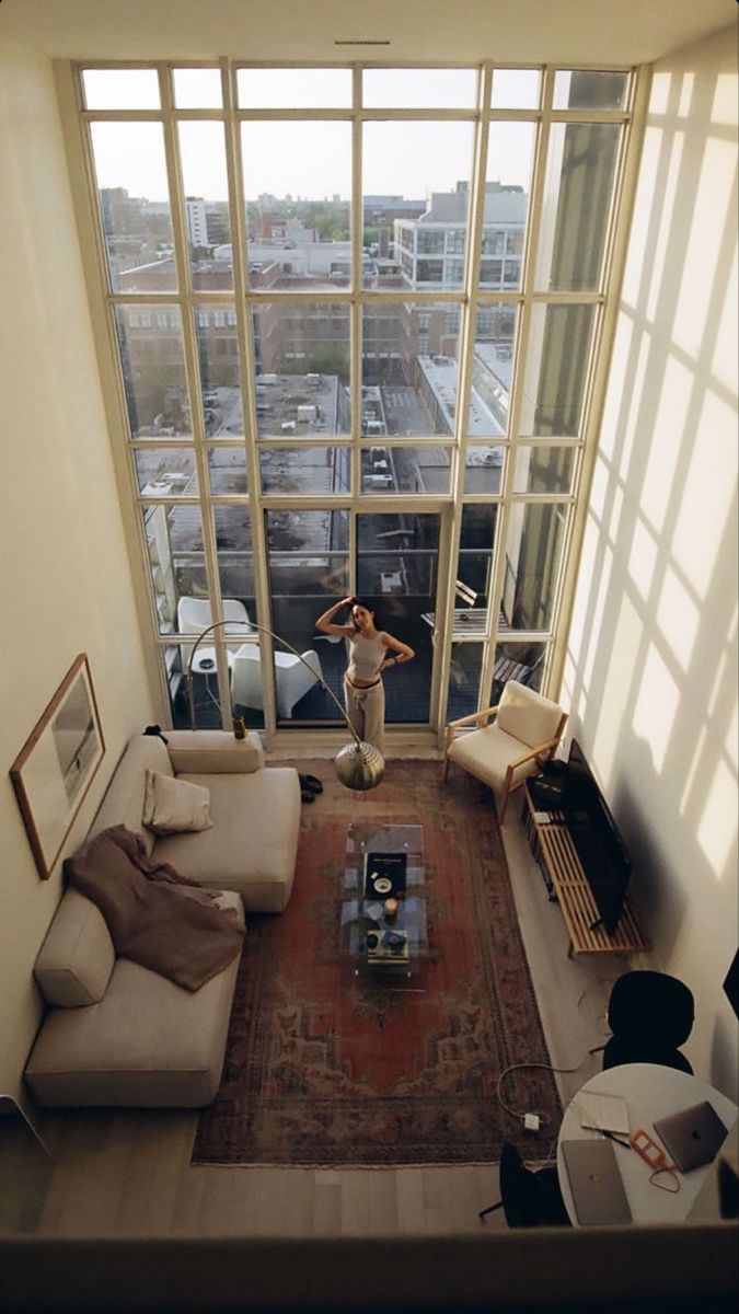 a highangle, full shot of a loft apartment with floortoceiling windows offering a panoramic city view. sunlight streams into the open space, illuminating a stylishly furnished living room.  a woman stands near the windows, and the room features a beige sofa, armchairs, a coffee table, and a rug.  a laptop sits on a nearby table suggesting a home office setup. the overall aesthetic is minimalist, modern, and serene.