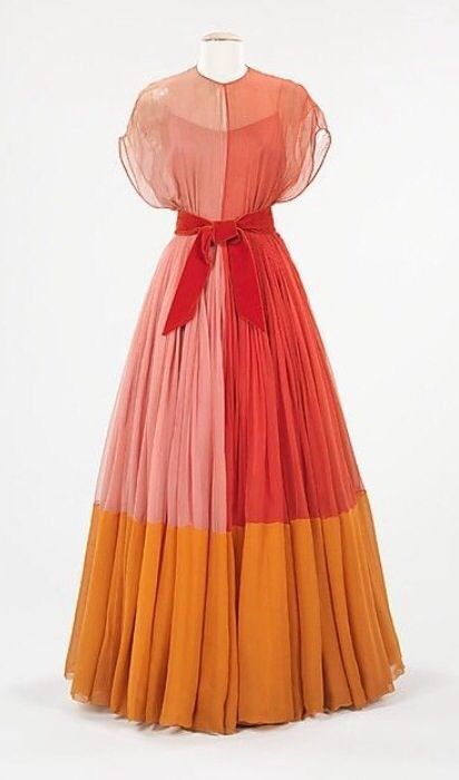 a floorlength chiffon dress is displayed on a mannequin. the dress has a color block design with sections of pink, red, and orange, separated by a red velvet belt. the top portion of the dress is sheer.