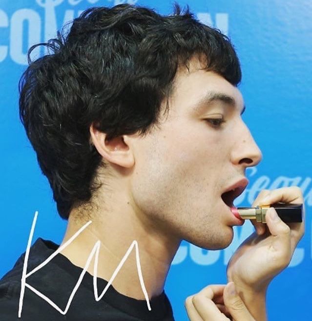 a profile view of ezra miller applying lipstick. they are holding a black and gold lipstick tube in their right hand and applying it to their open mouth. ezra miller has dark, wavy hair and is wearing a black shirt. the background is blue with faint text. a white "km" is written on the left side of the image.