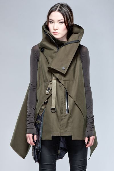 a young woman with long dark hair is posing for the camera in a studio. she is wearing a long, green jacket with a large collar, which is styled to look like a vest. the jacket is decorated with straps and buckles. the woman is also wearing black pants.